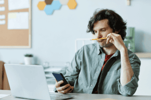 man holds a pencil to his upper lip while looking at his phone sitting at a desk and questions himself do i have adhd
