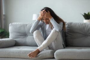 a struggling woman sits on her couch holding her hands to her head and thinking about her trauma responses