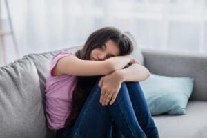 an upset teen girl sits on a couch with her legs hugged into her chest wondering if shes experiencing signs of depression in teens