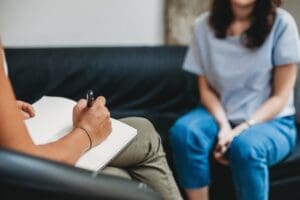 person sits on a couch during therapy session while therapist takes notes in a notepad